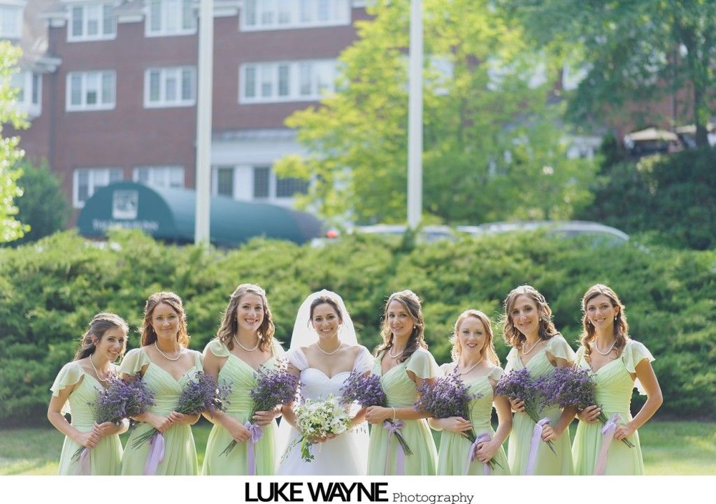 Bridesmaids in green dresses with lavender bouquets pose with bride in front of brick building.