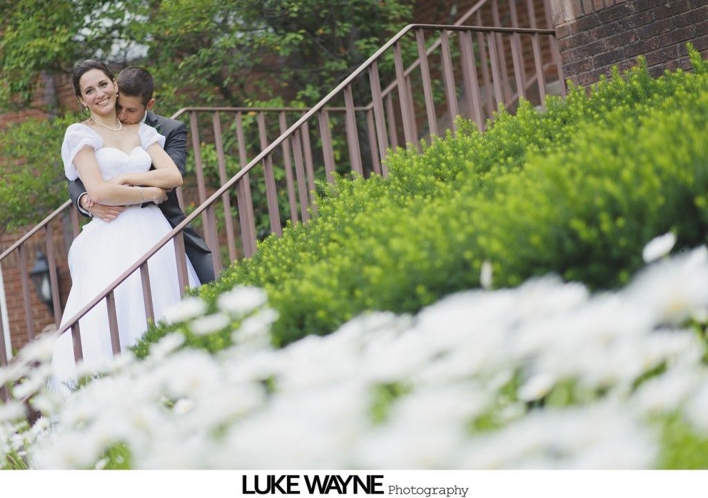 Bride and groom embrace on steps, next to green bushes and white flowers.