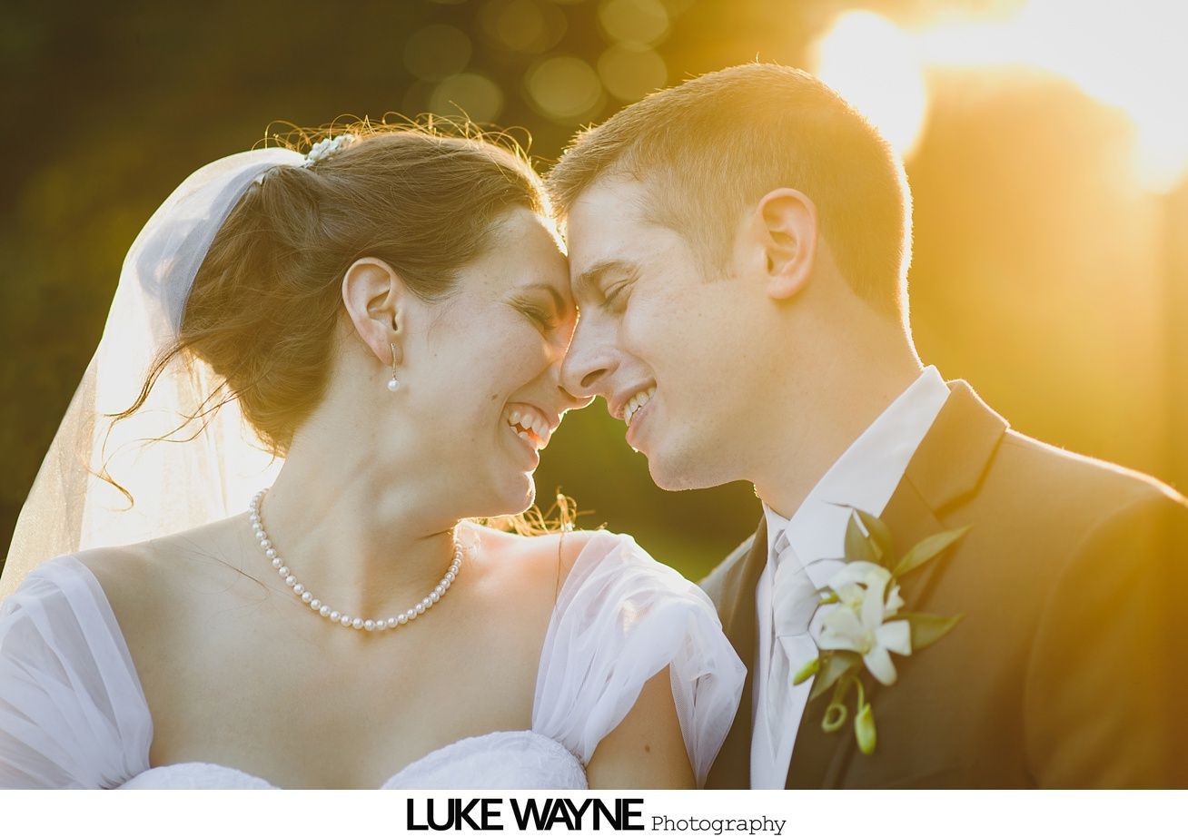 Bride and groom smiling, forehead to forehead, bathed in golden sunlight. Bride wears veil, necklace; groom in suit.