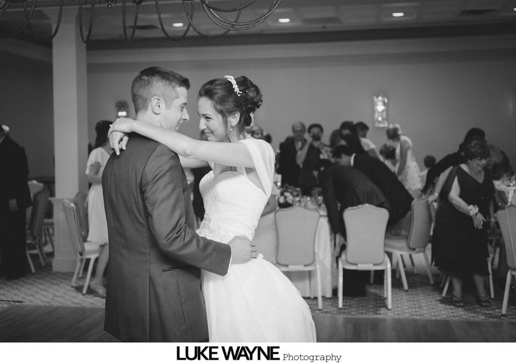 Bride and groom dance at a wedding reception; guests watch from tables in a ballroom.
