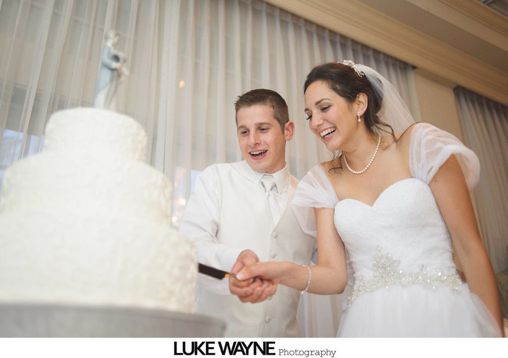 Bride and groom cutting wedding cake. They are smiling. Indoor setting.