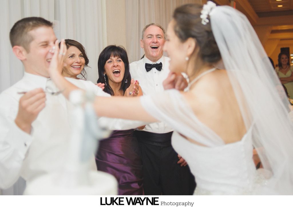 Bride and groom playfully smearing cake on each other during a wedding reception, guests laugh.
