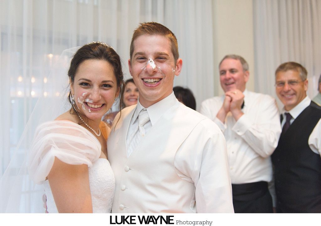 Bride and groom with cake on their faces smiling at wedding. Guests in background.