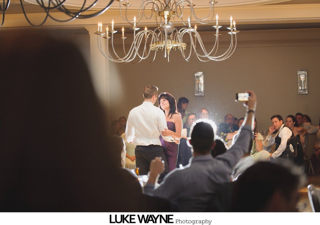 Couple dances under a chandelier as guests watch and take photos in a brightly lit room.