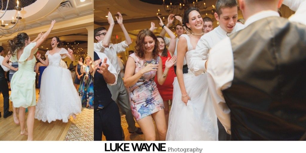 Wedding guests dancing joyfully on a wooden floor in a ballroom. The bride in a white dress is in the center.