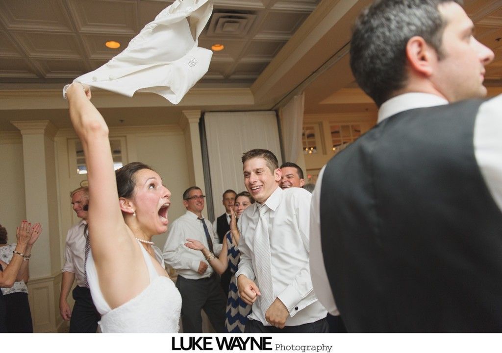 Bride throws shirt in air during wedding reception. Dancing guests, indoors.