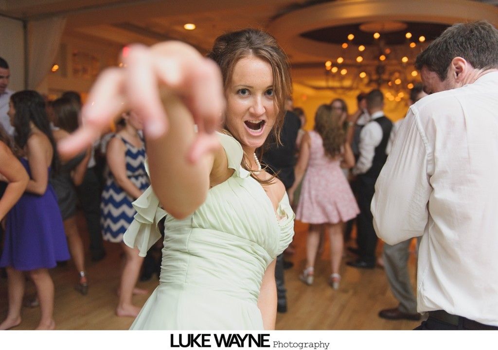 Woman in a light green dress reaches toward the camera on a dance floor, people dancing in the background.
