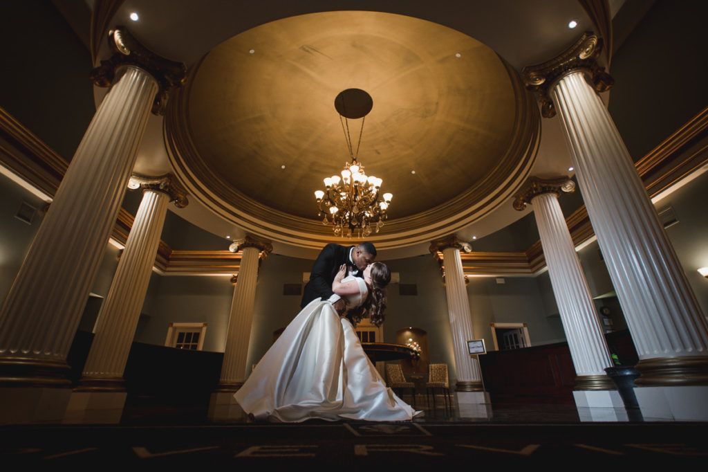 Couple kissing under a domed ceiling with a chandelier, flanked by columns, during a wedding.