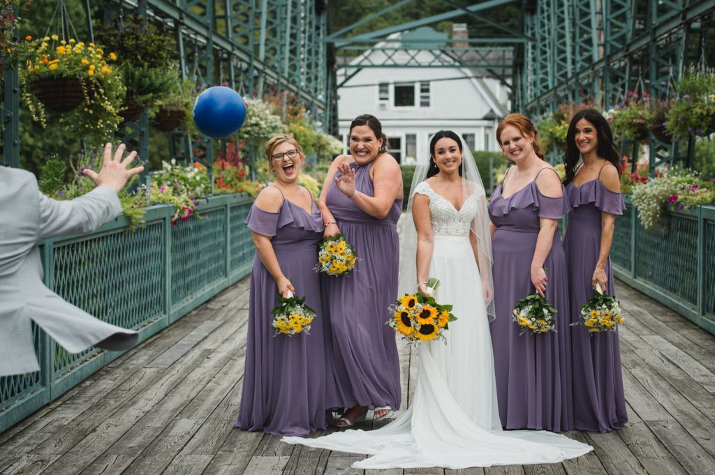 Bride and bridesmaids in purple dresses on bridge; man throws balloon.