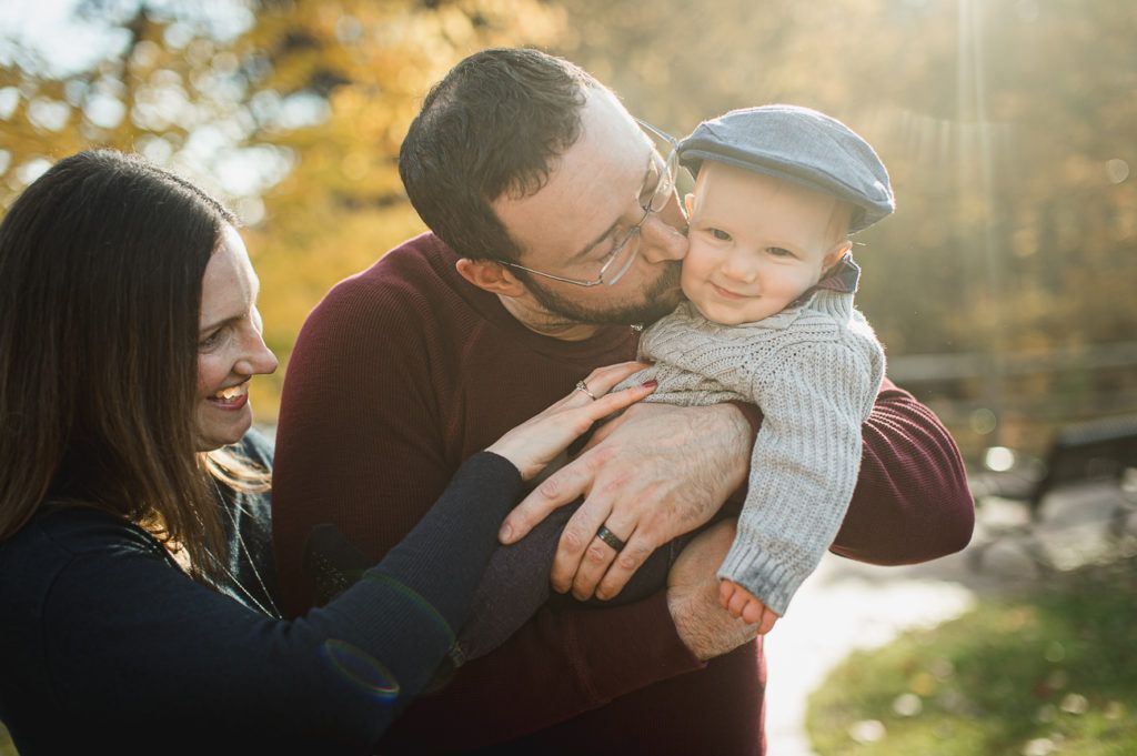 Parents smiling, kissing baby wearing a cap. Bright outdoor setting.