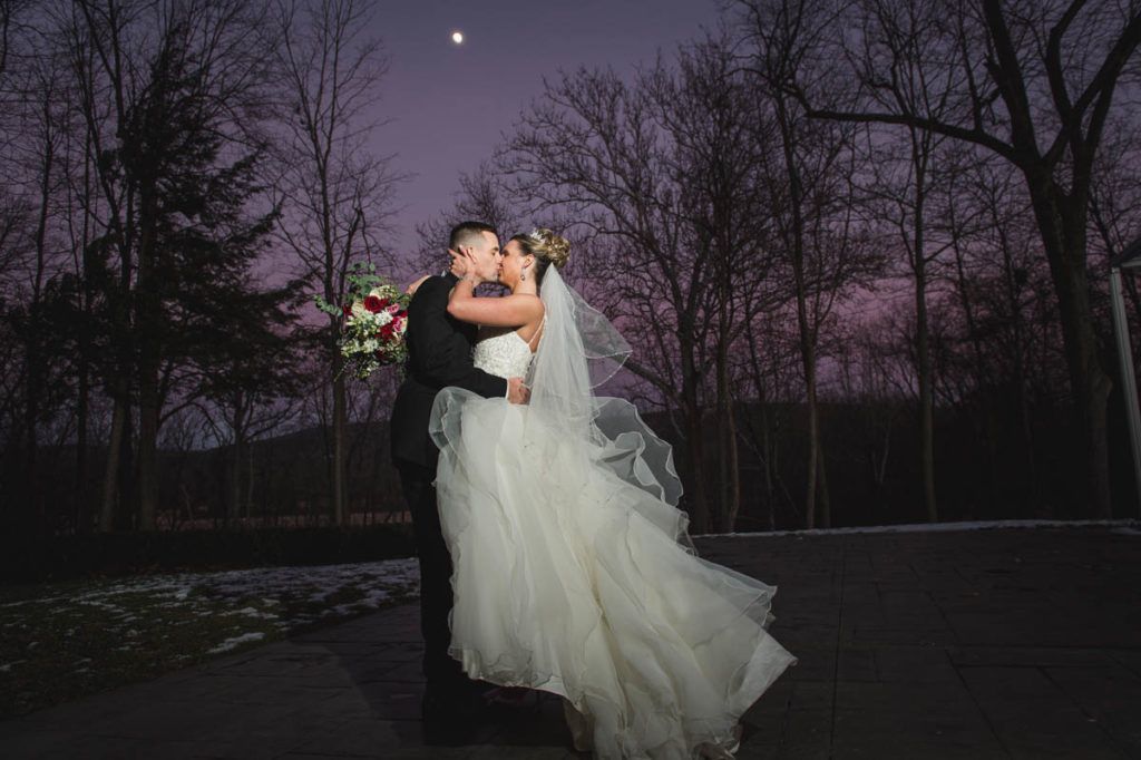Bride and groom embrace, kissing, in front of a tree-lined backdrop at dusk.