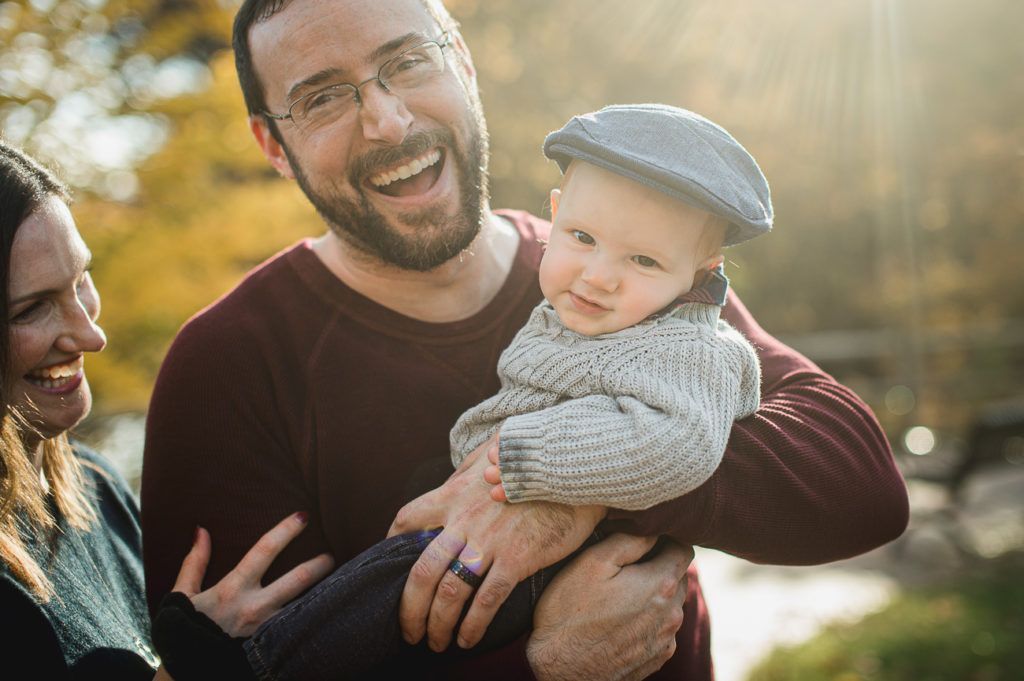 Man holding baby wearing hat, laughing. Woman partially visible, blurred autumn background.