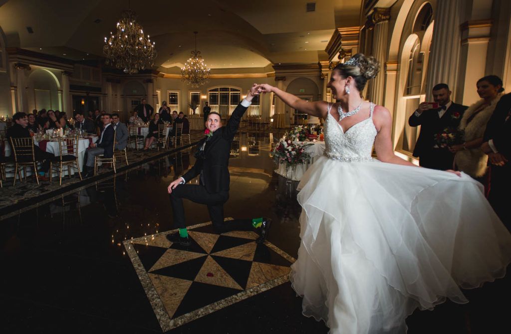 Bride twirls as groom kneels on a checkered dance floor in a ballroom, guests watching.