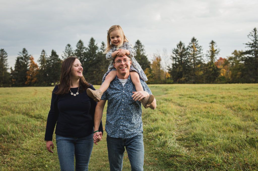 Family in a field: Father carrying a child on his shoulders, mother walking beside them, smiles, cloudy sky.