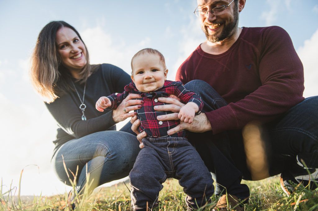 Parents supporting a baby learning to walk outdoors; sunny day.