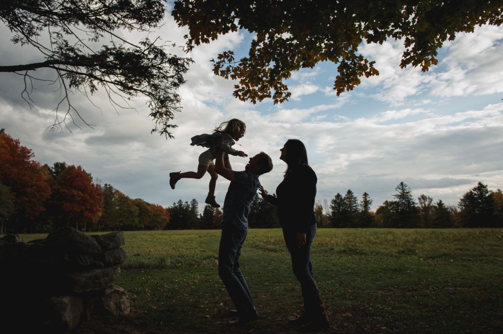 A parent throws a child into the air as the other parent looks on in a field beneath a tree.