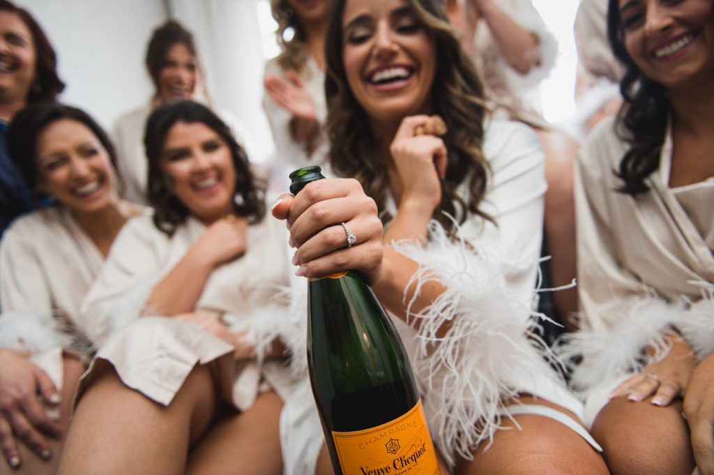 Group of women in robes laughing, opening champagne bottle indoors.