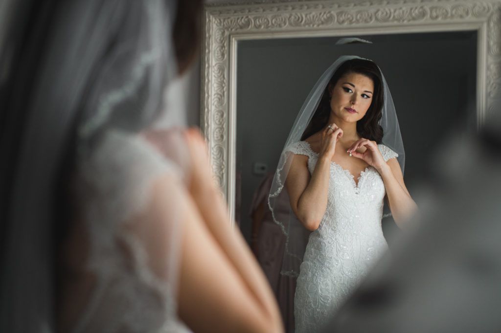 Bride in a white dress and veil adjusts necklace in front of a mirror.