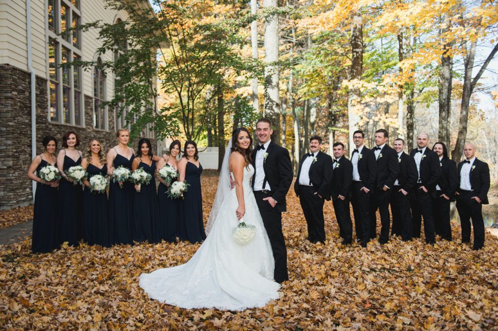 Wedding party poses outdoors: bride and groom in front, bridesmaids in navy, groomsmen in black, surrounded by autumn leaves.