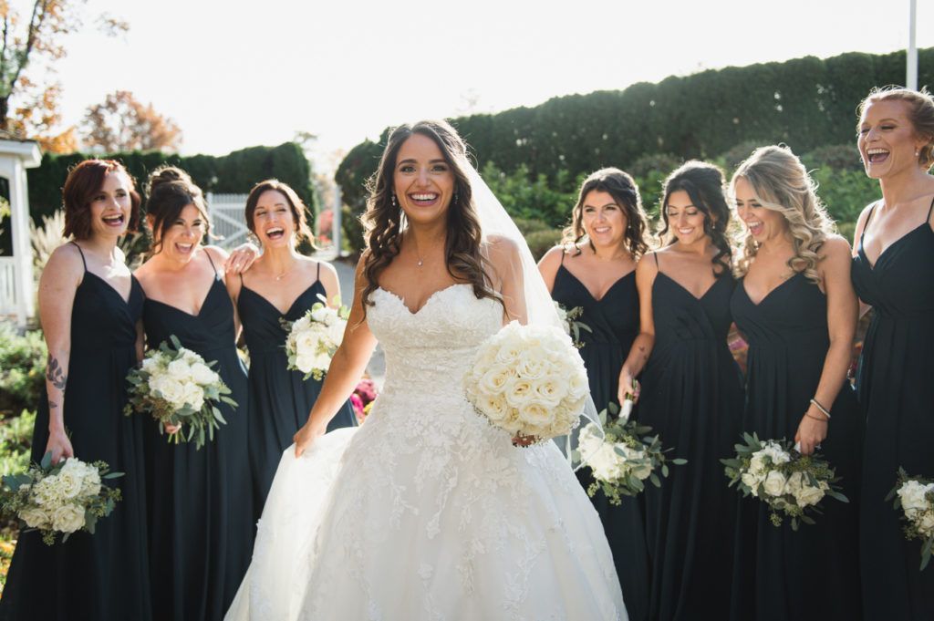 Bride in a white gown smiles with bridesmaids in navy dresses holding bouquets; outdoor setting.