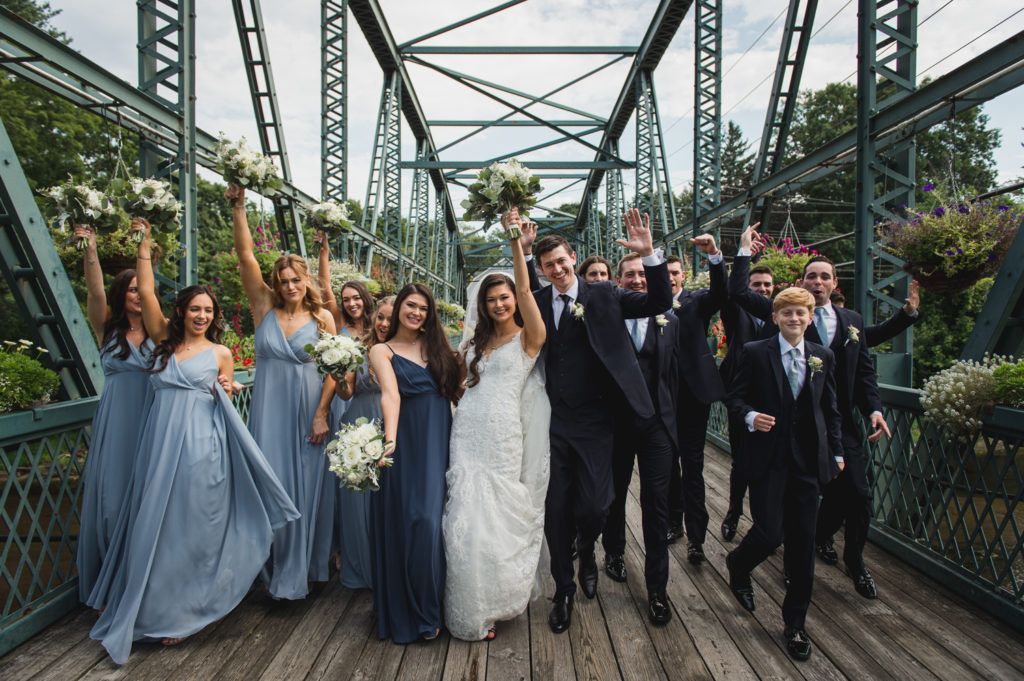 Wedding party on a green bridge, cheering with bouquets raised. Bride and groom in center, bridesmaids in blue dresses, groomsmen in suits.