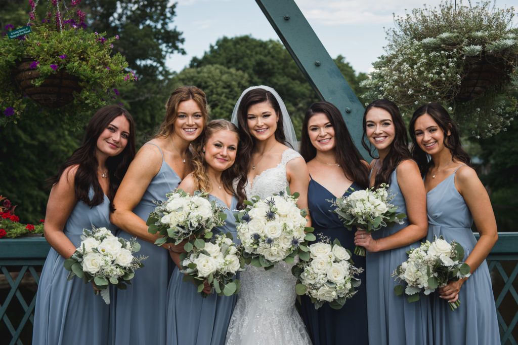 Bride with bridesmaids, smiling, holding bouquets on a bridge. Dresses are varying shades of blue.