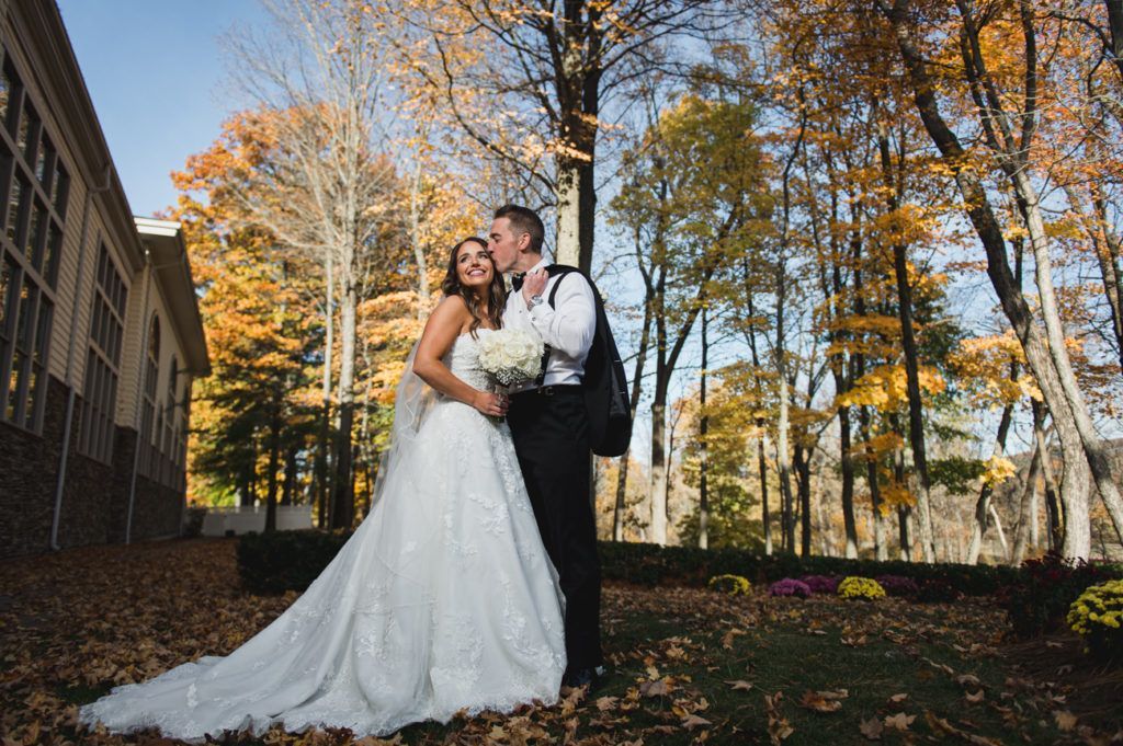 Bride and groom share a kiss outdoors. Autumn leaves frame them; he wears a tux, she a wedding dress.