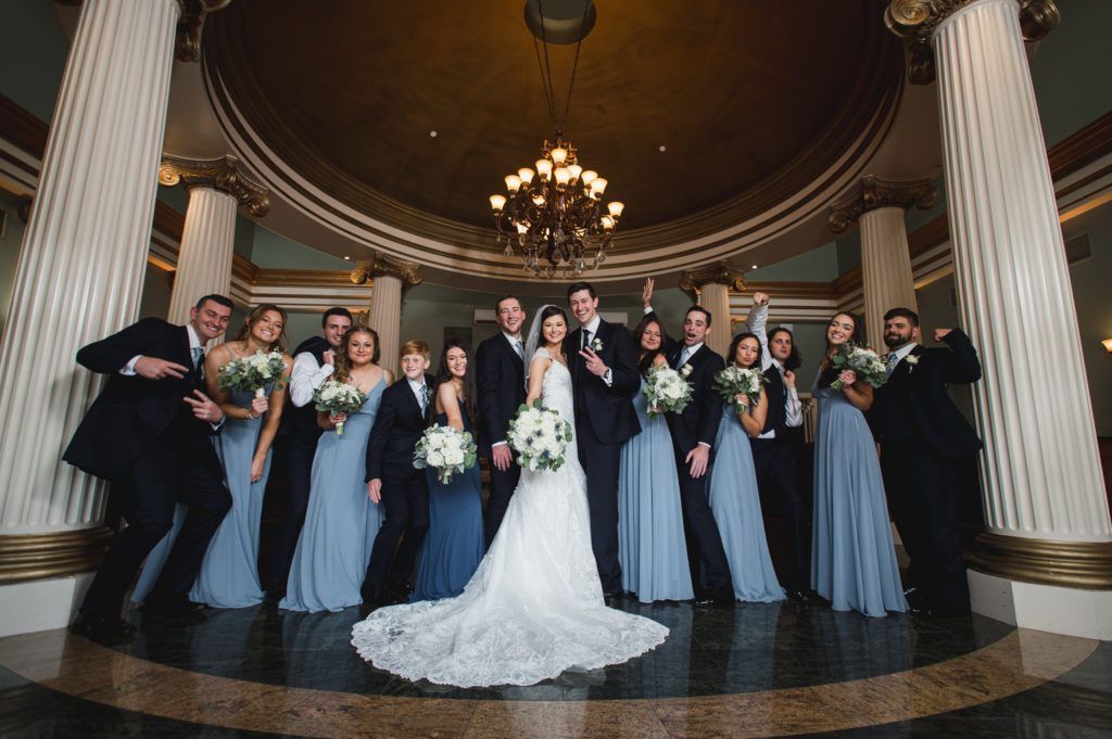 Wedding party poses in front of grand columns and a chandelier. The bride and groom are center, with bridesmaids in blue.