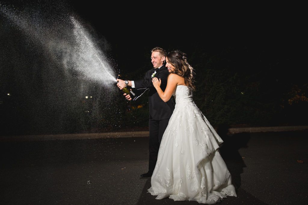 Newly married couple spraying champagne at night; woman in white gown, man in tuxedo.