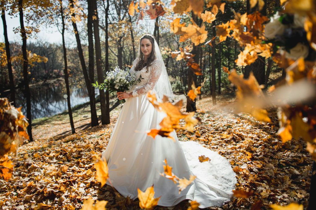 Bride in a white gown and veil holds a bouquet, surrounded by falling orange leaves in a forest.
