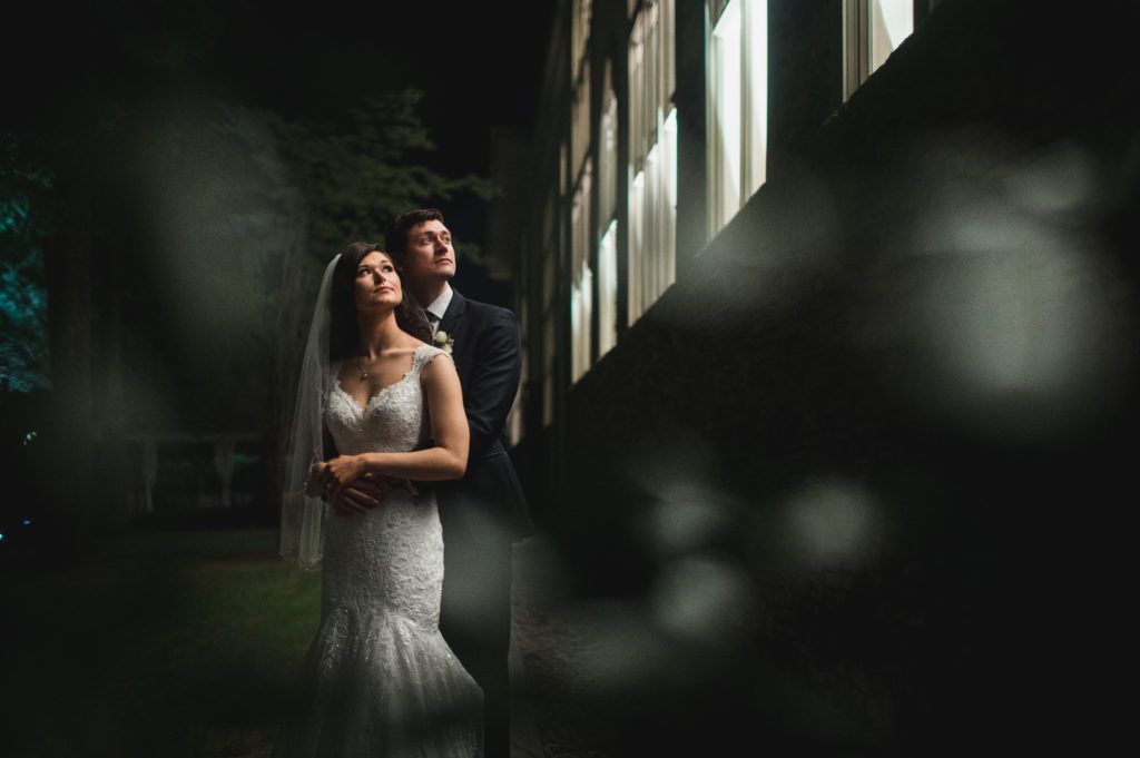 Bride and groom in wedding attire look upwards near illuminated windows at night.