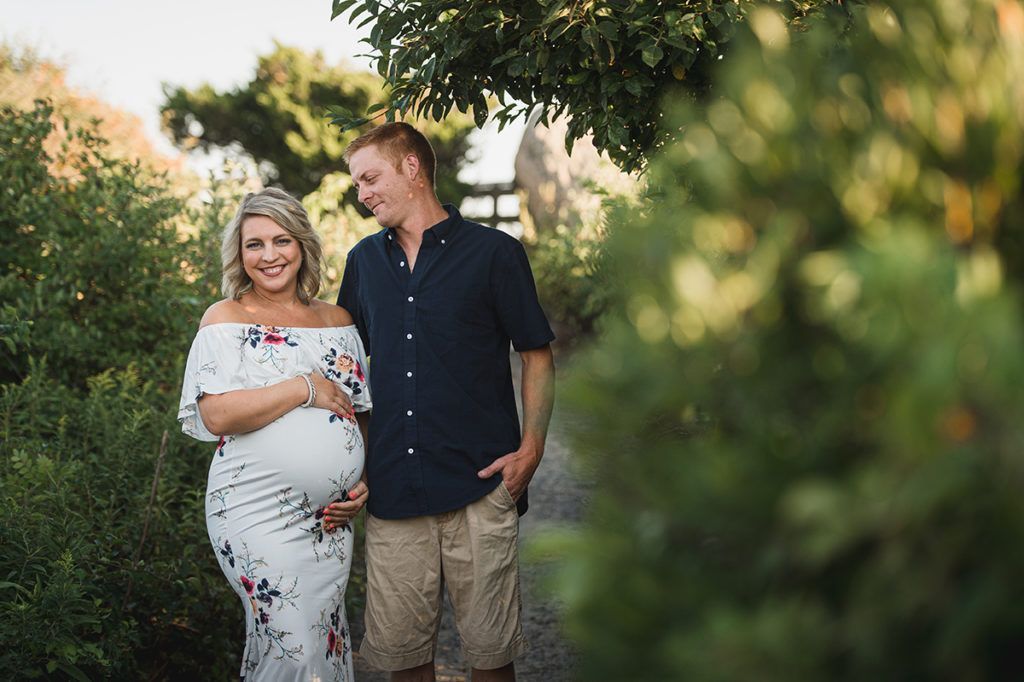 Pregnant woman in floral dress and man in shorts smile, standing outdoors.