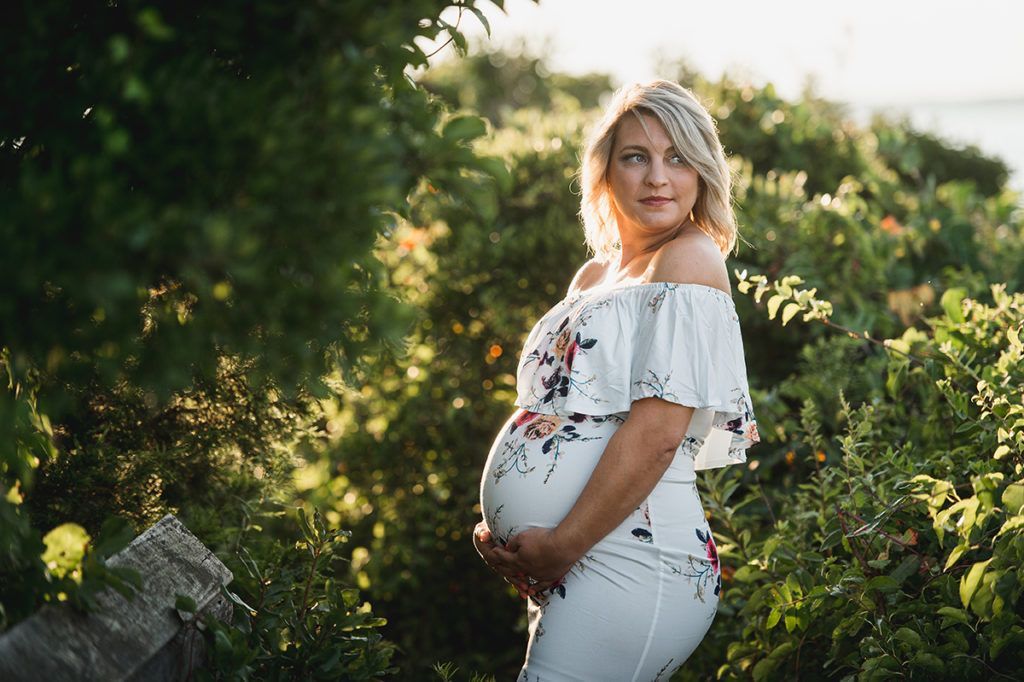Pregnant woman in white floral dress, outdoors, holding belly, smiling.