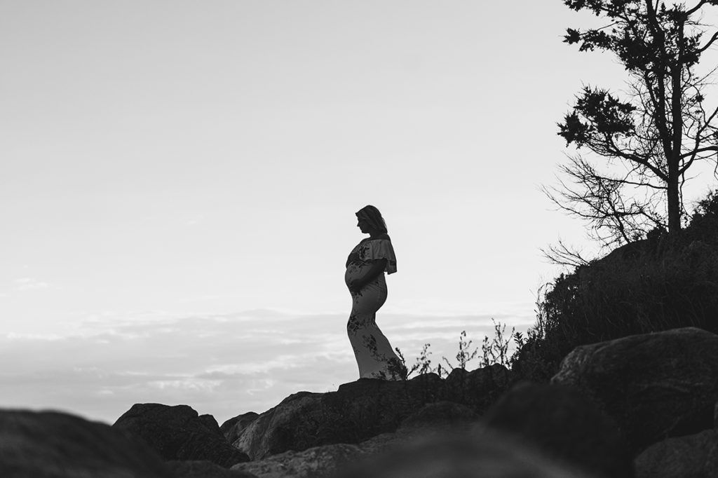 Pregnant person standing on rocks, ocean in the background, backlit, silhouette.