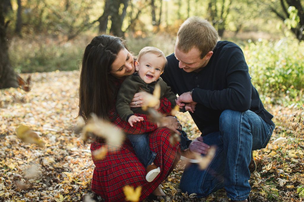 Family of three in fall leaves: parents smiling at baby, throwing leaves in forest.
