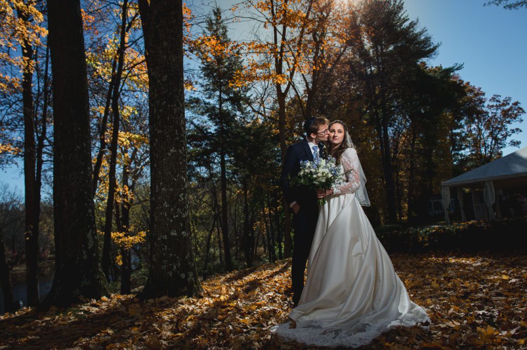 Bride and groom pose in autumn woods; the bride wears a white dress and holds a bouquet. Sunlight streams through trees.