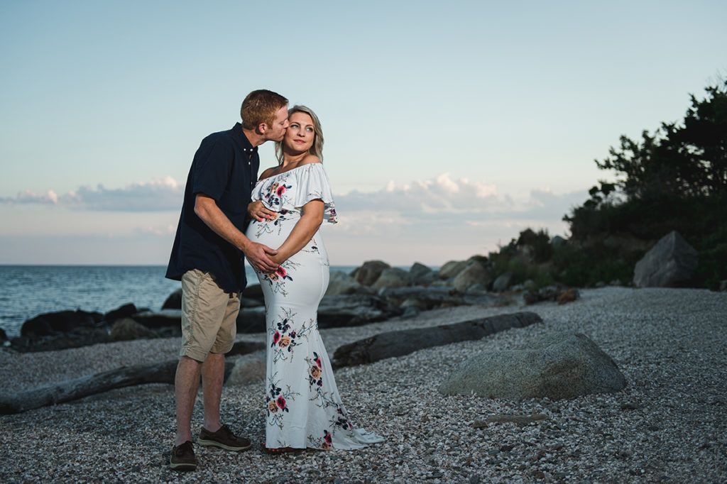 Man kisses pregnant woman's cheek on a beach at sunset, both holding her belly.