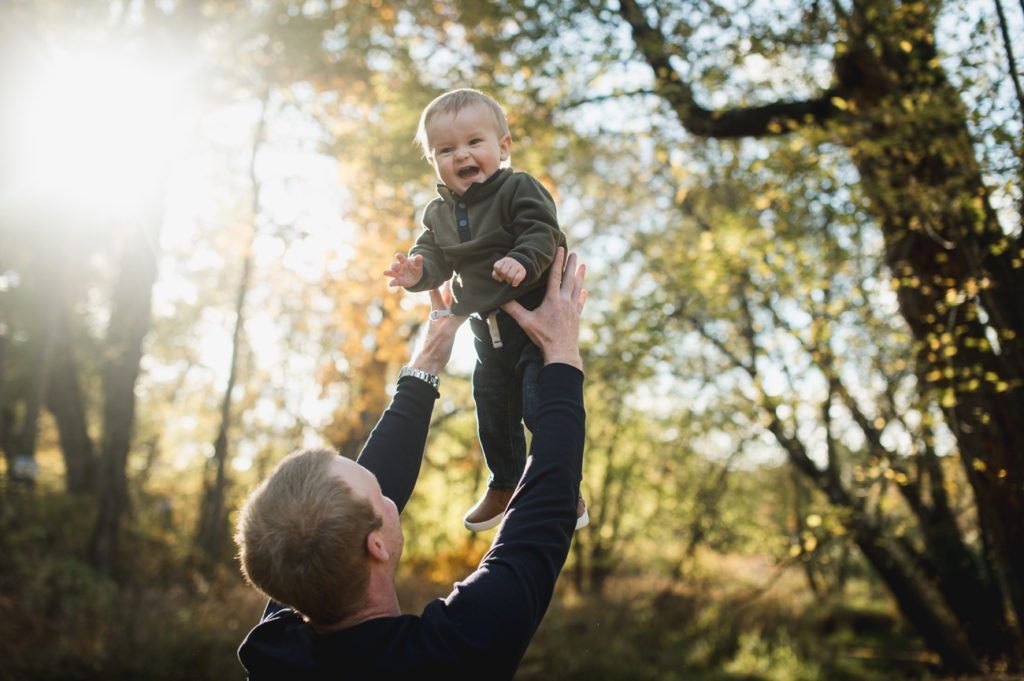 Father throwing a smiling baby boy up in the air outdoors with trees in the background, sunlit scene.