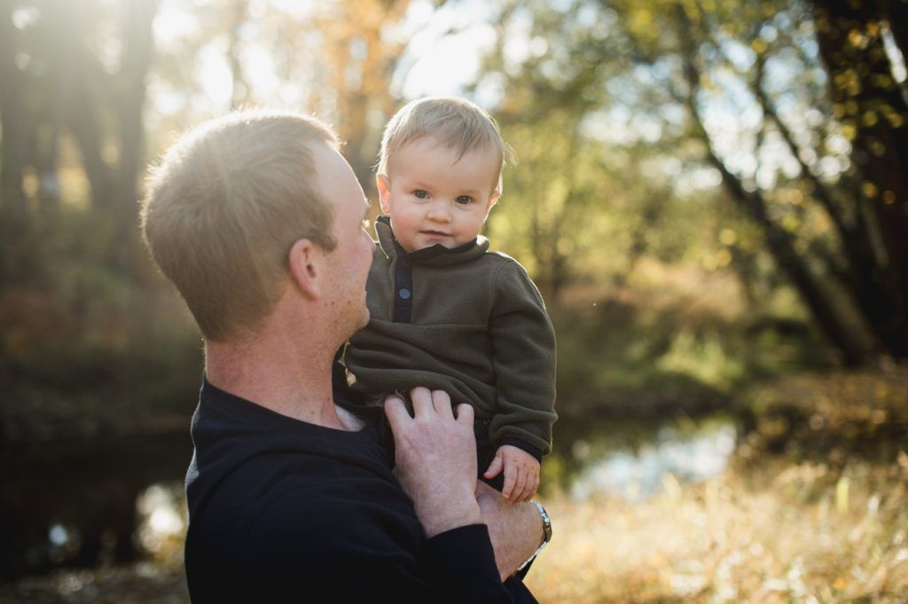 Man holding a baby outdoors near water, both looking at the camera. Sunlight streams through the trees.