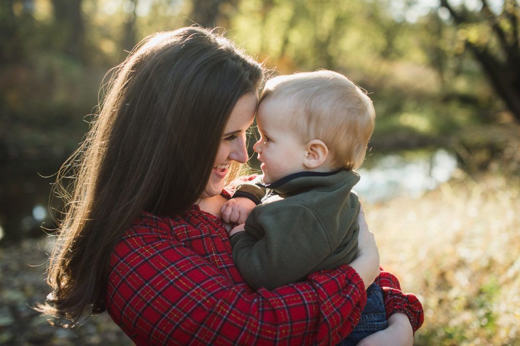 Woman in red plaid shirt holds a baby, face to face outdoors. Backlit by sun.