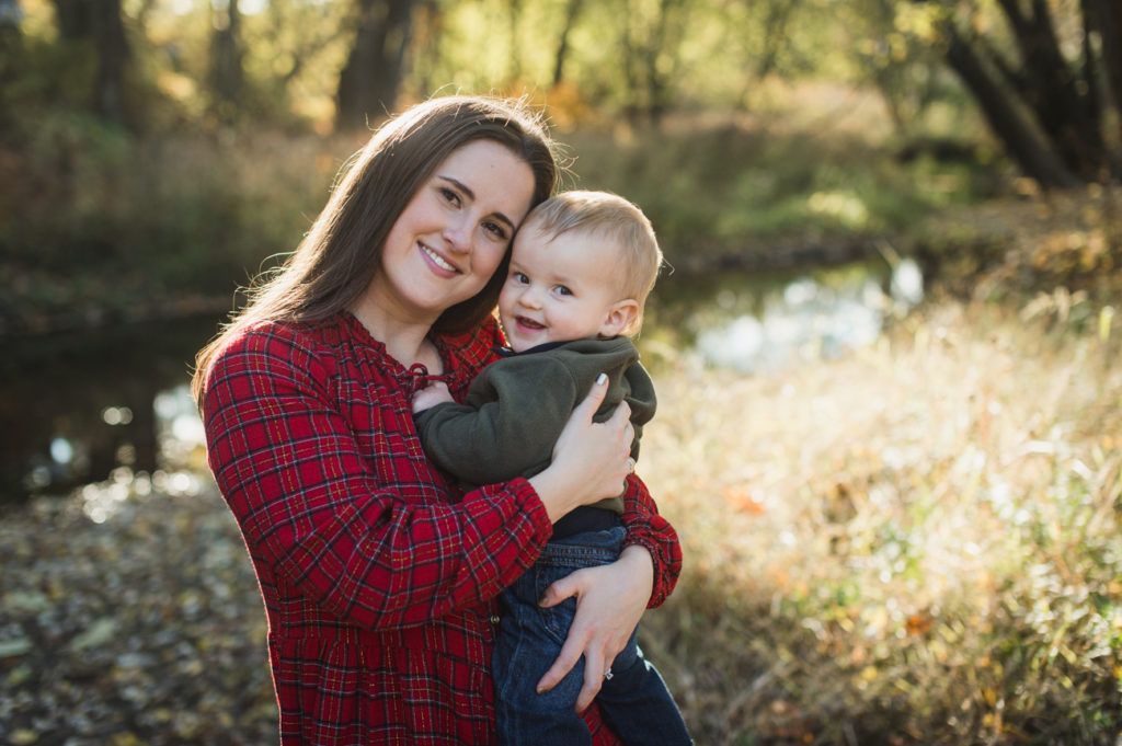 Woman in red sweater holds a smiling child in an outdoor setting with sunlight, trees, and a stream.