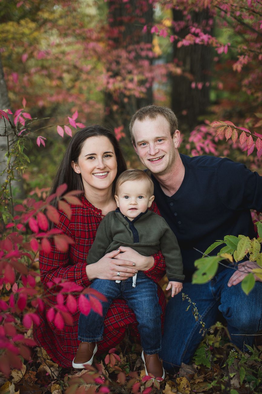 Family of three smiles amongst red foliage.