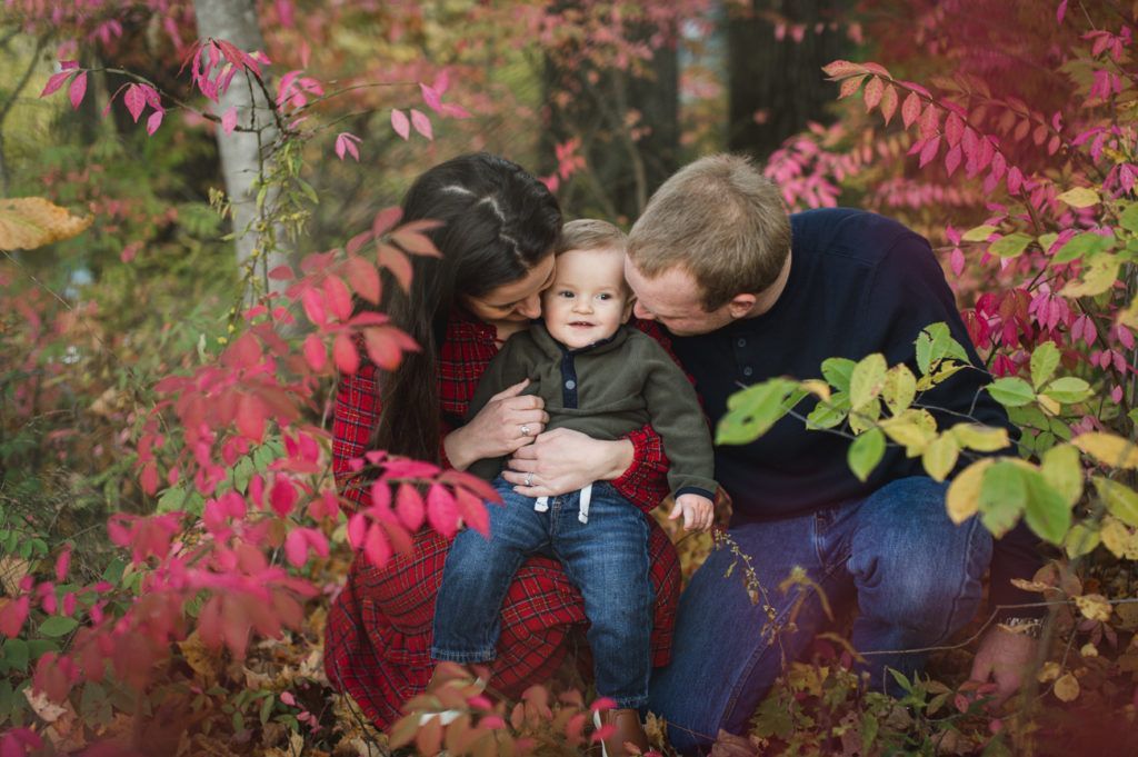 Family embraces outdoors, amidst fall foliage, red and yellow leaves. Parents kiss child on each cheek.