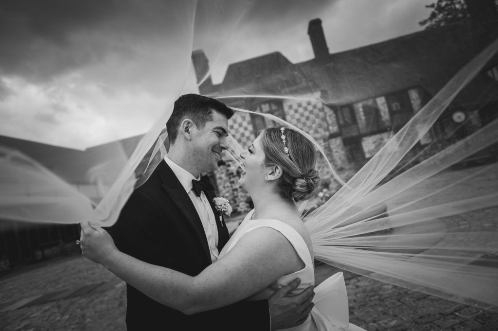 Bride and groom embrace, wedding veil flowing. Building in background, black and white photo.