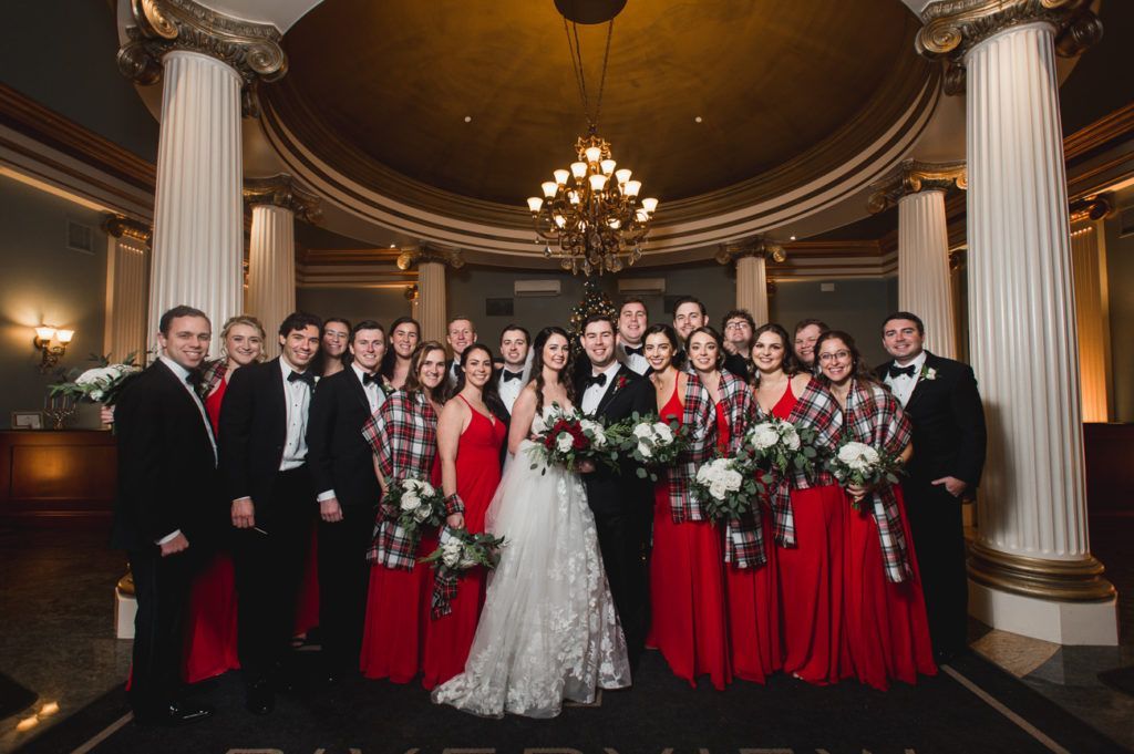 Wedding party posing in a grand hall; bride and bridesmaids in red, groomsmen in black, tartan shawls.