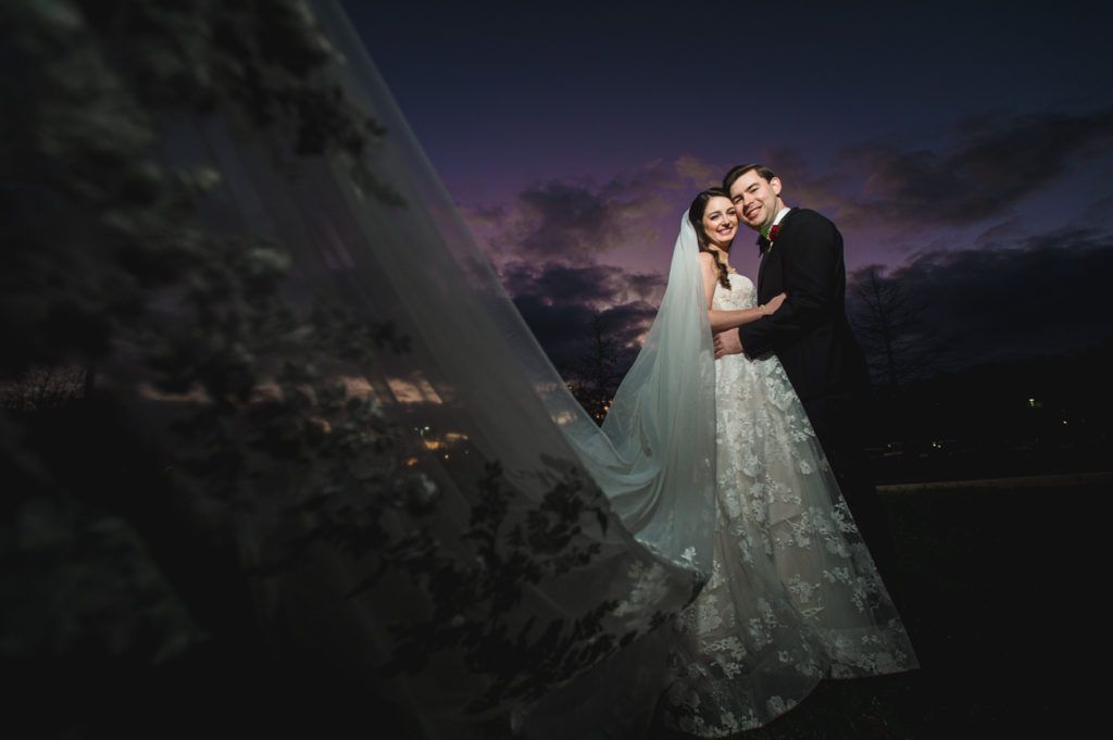 Bride and groom embrace outdoors at sunset, veil billowing in the wind, a night sky backdrop.