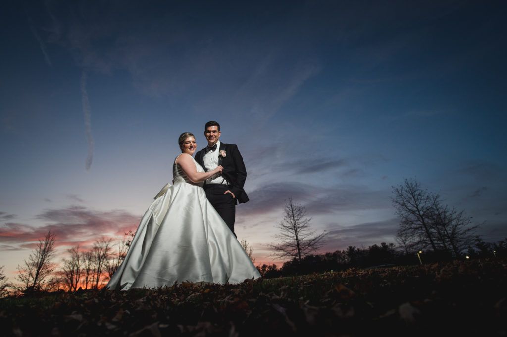 Bride and groom pose on hill at sunset; woman in white dress, man in tuxedo.