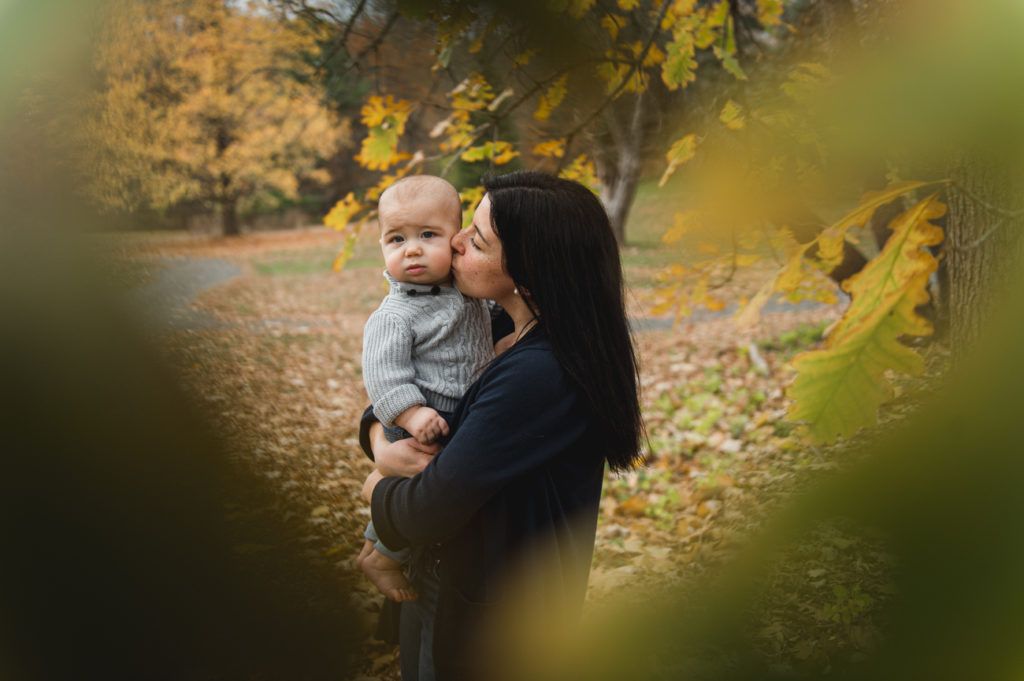 Woman kissing a baby held in her arms in a park with autumn foliage.