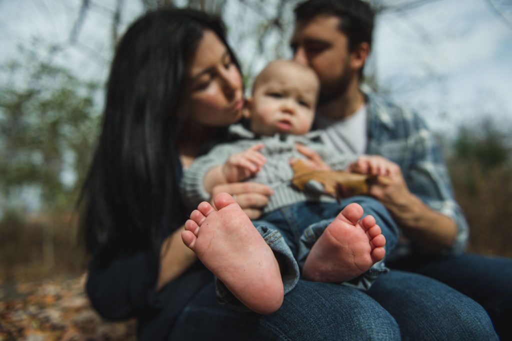 Family cuddles outdoors; parents kissing baby held on lap.