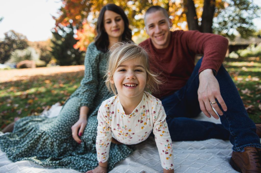 Family of three smiling outdoors, autumn setting; girl in front, parents behind on blanket.